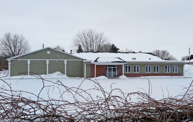Color photograph taken in winter, long shot of facade of large rectangular building with flat roof and beige vinyl siding with a few windows, next to the front door, a large brown cross, in the foreground, a fence filled with vines.