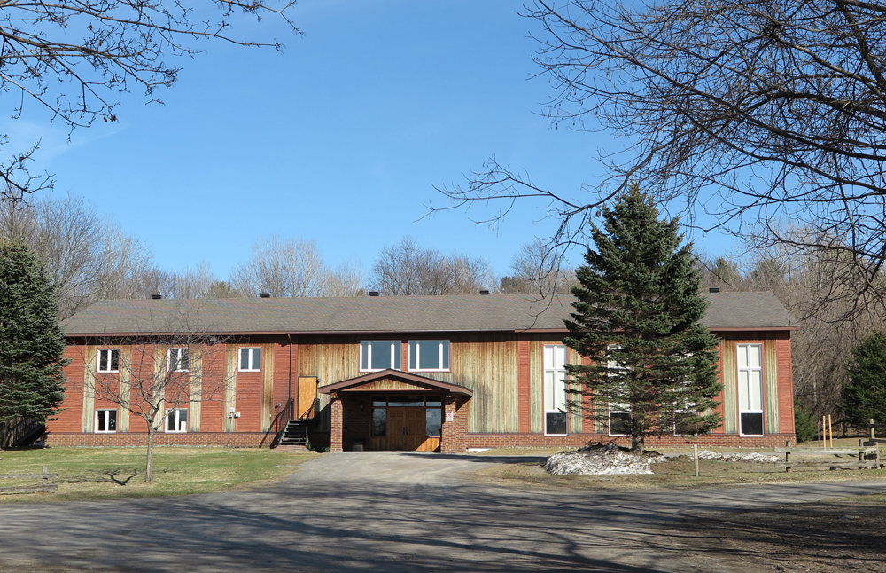 Color photograph, long shot of façade of a large rectangular wood building with many square windows, in the foreground, a large paved road leads to a wood main door.