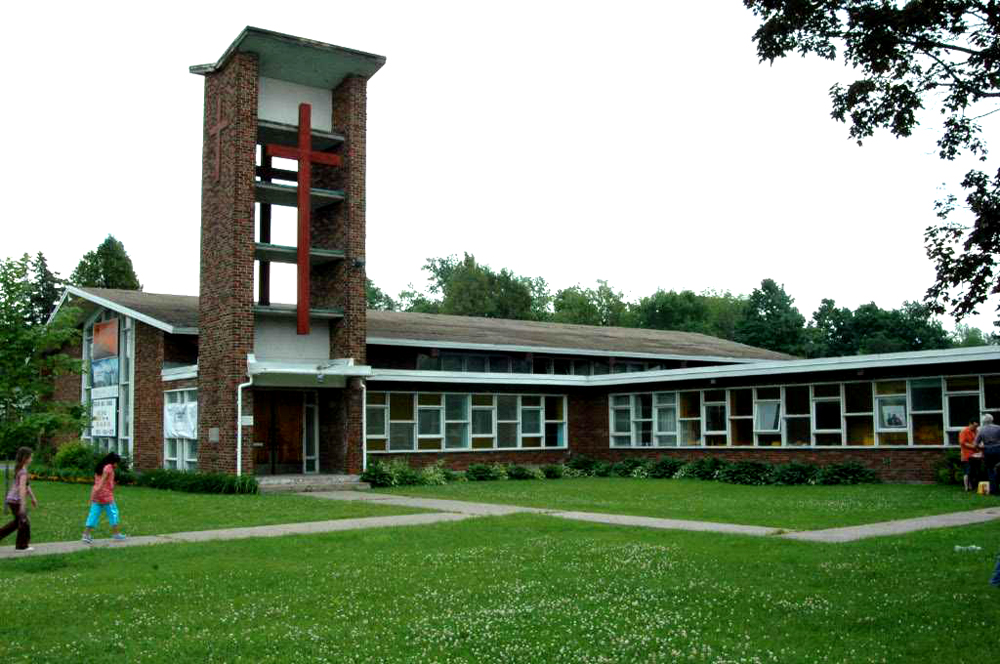 Color photograph, long shot of a brown brick building made of two sections, to the left, the church with sloped roof and open tower topped with a large cross, to the right, a building with a flat roof with many windows, in the foreground, children walking on a sidewalk.