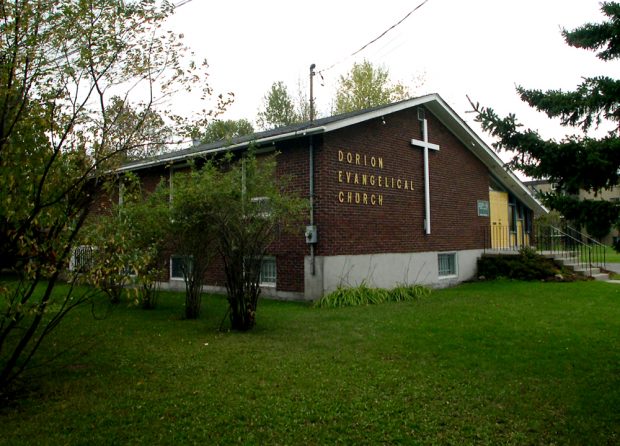 Color photograph, long shot of side-view of a brown brick building with a sloped roof, on the façade, a large white cross next to which is written: Dorion Evangelical Church.