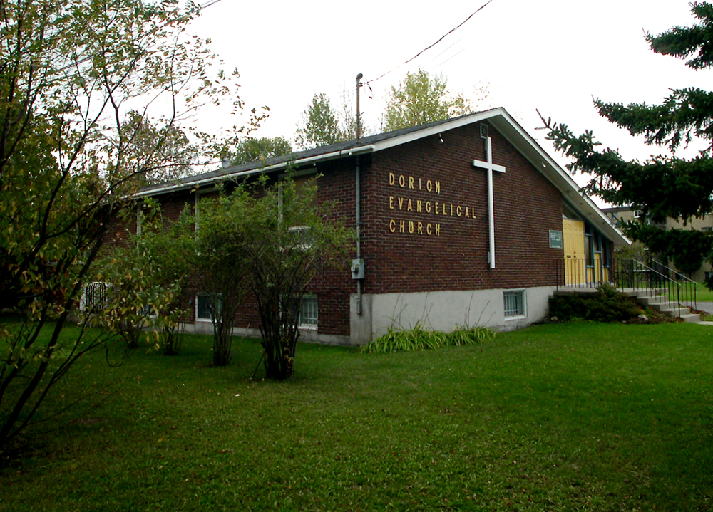 Color photograph, long shot of side-view of a brown brick building with a sloped roof, on the façade, a large white cross next to which is written: Dorion Evangelical Church.