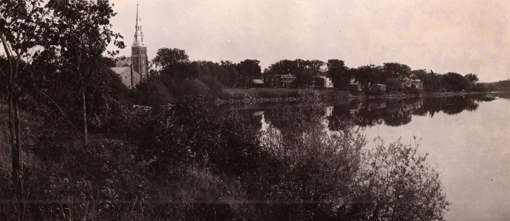Old black and white photograph, large body of water, trees and vegetation in the foreground, a church steeple and a row of small houses in the background.