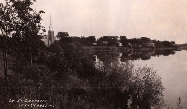 Old black and white photograph, large body of water, trees and vegetation in the foreground, a church steeple and a row of small houses in the background.