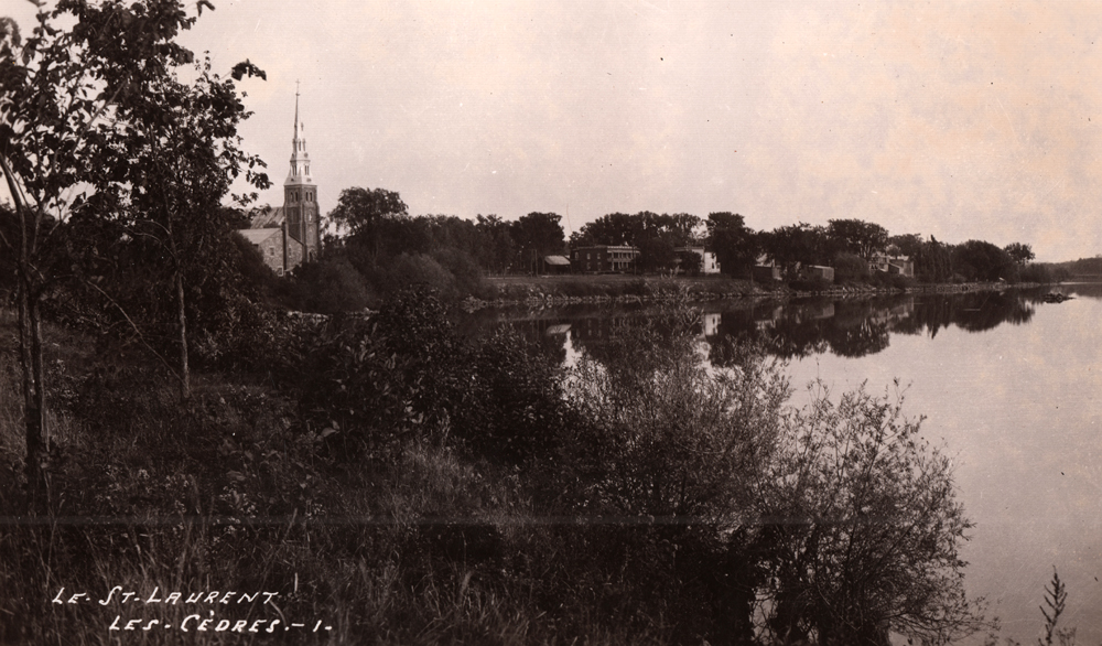 Old black and white photograph, large body of water, trees and vegetation in the foreground, a church steeple and a row of small houses in the background.