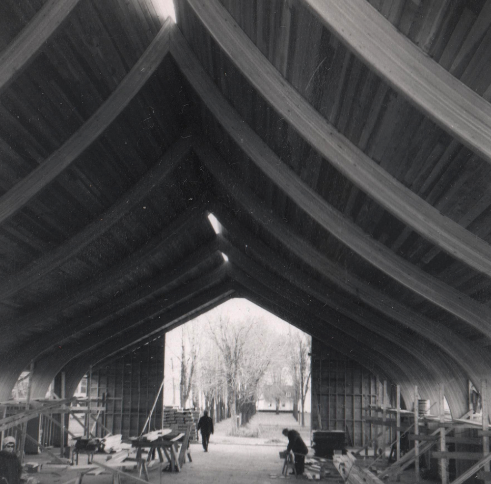 Black and white photograph, close-up of the wood framework of a building under construction, the rear wall is unfinished, inside are building materials and workers, in the foreground on the left, a woman looks toward the camera.