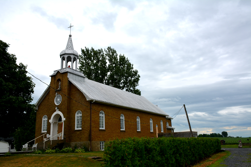 Color photograph, red brick church with steeple and statue in an alcove, on the church porch is garden furniture, on the right of the building, a boarded up window and a concrete slab annex, fields in the background.