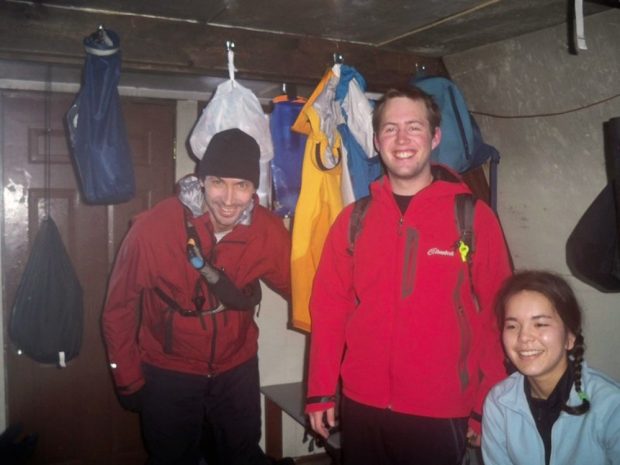 Three members pose for the photo, two men wearing red jackets and black pants are standing and a women in a sky blue fleece is sitting near the front entrance. Behind the men standing, bags and jackets hang from the support beam holding up one of the Hut arches.