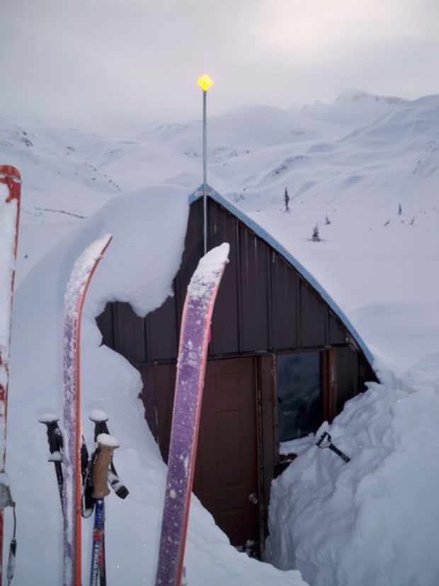 Purple and pink ski tips and ski poles stuck in the snow to the left of the partially dug out front entrance and window of the Gothic Arch Hut.