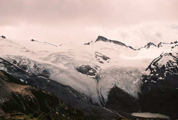 A massive glacier covers the slope and obscures the peaks above. The creep of the glacier extends to the hanging alpine valley floor. Grey clouds swirl above the high peaks.
