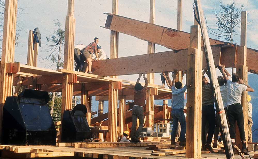 Several members work at construction, raising a large wooden floor joist and maneuvering it into position.