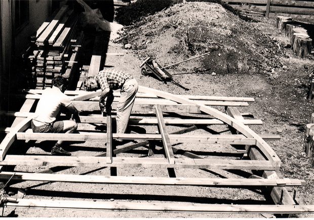The arch frame sits off the ground supported by two other pieces of wood. Two men are hunched over working on pieces of the arch on the left hand side. Other lumber sits stacked neatly behind next to a house.