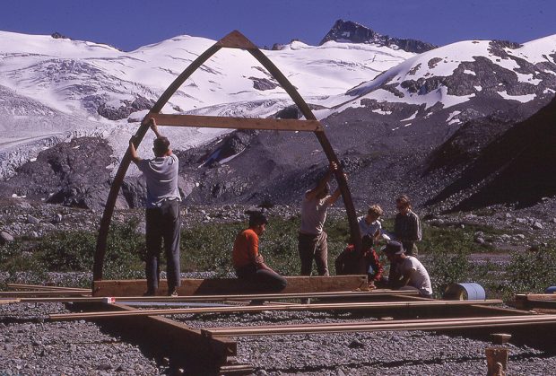 The foundations have been built and several pieces of the floor stretch across the width above the rocky ground below. Two men hold up the first wooden arch, another sits in the middle of the arch while another group work to attach the arch to the foundation.