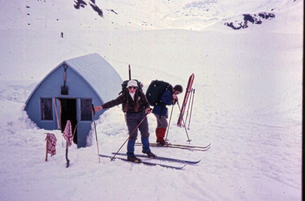 A women wearing a white hat, sunglasses, skis and has long pigtails stands next to a man with a black backpack and a blue jacket stepping into his skis. The Hut sits behind and the front entrance has been dug free of snow.