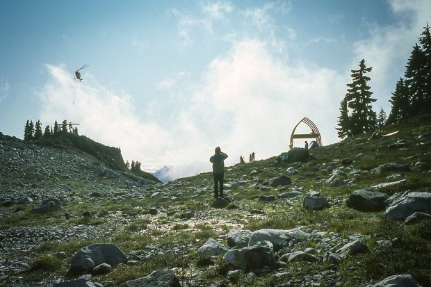 A helicopter carrying a load of supplies flies on the far left; in front another member is taking a photograph and off In the distance is the bare arched frame structure and other UBC-VOC members.