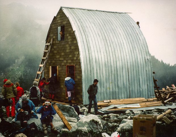 The Hut structure is completed with aluminum siding. A man looks out the top floor window above the main entryway. Other members are seen working on the front end wall and in front of the entrance to the Hut.