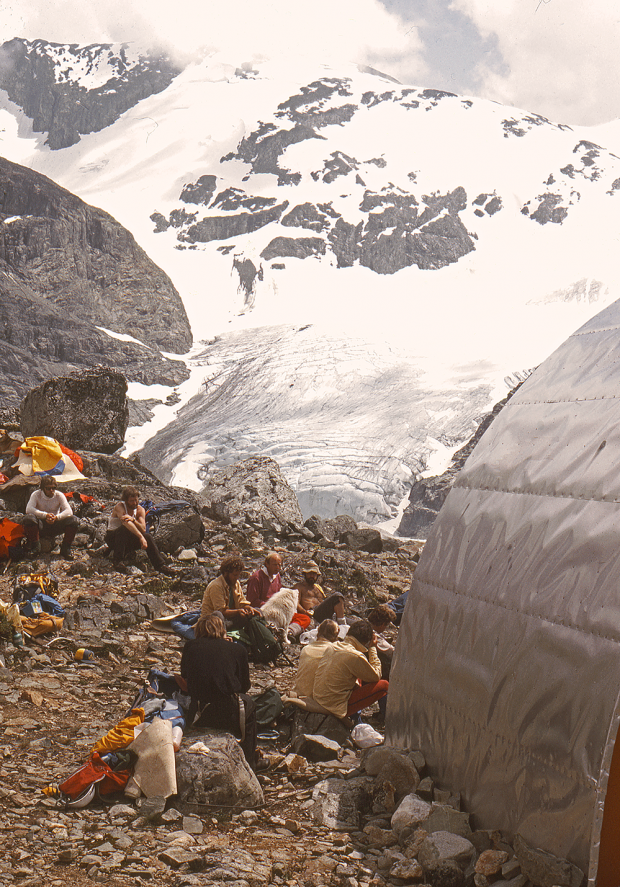 The side of the Wedgemount Lake Hut is in the foreground and a group of people sitting on rocks outside the hut. Wedge Mountain and glacier fill the background.
