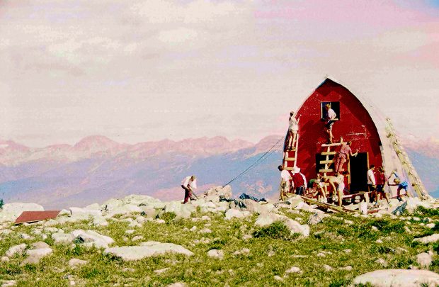 The Hut Structure has been built and UBC-VOC members are working on painting the front end-wall a bright red colour. Other members are busy working out front in the sunshine and bare mountain peaks can be seen off in the distance.