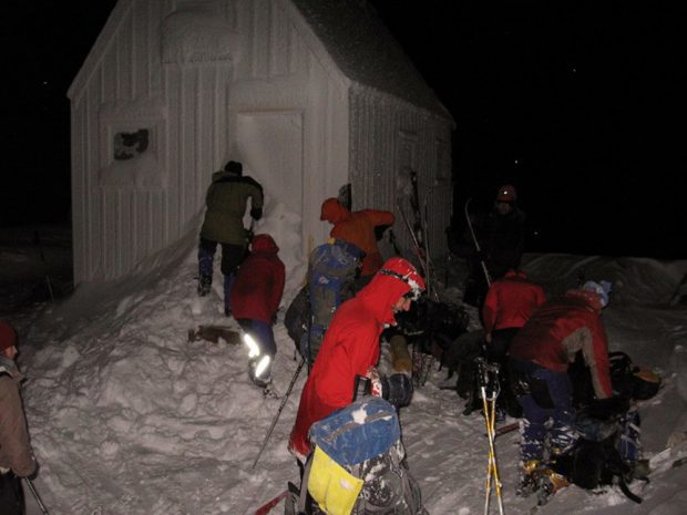 Three members near the front door of the hut are busy clearing the snow away. Off to their right a group of members are taking off ski equipment and organizing gear.