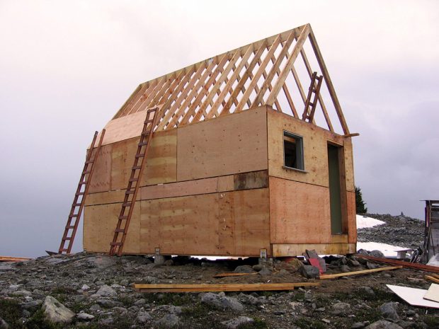 The wooden frame of the structure has been built and plywood has been applied to the exterior walls. The roof remains open to the elements and two empty wooden ladders lean up against the exterior.