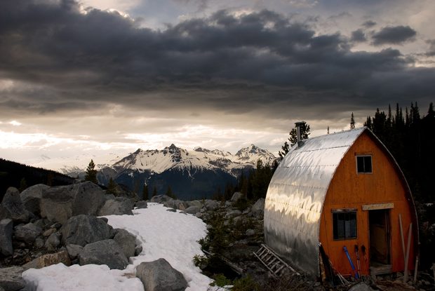 The Hut with its bright orange end wall and bright silver siding reflecting sunlight sits free of snow. The alpine meadow behind the hut is dotted with small boulders, evergreen trees and patches of snow. Snowy peaks are seen off in the distance.