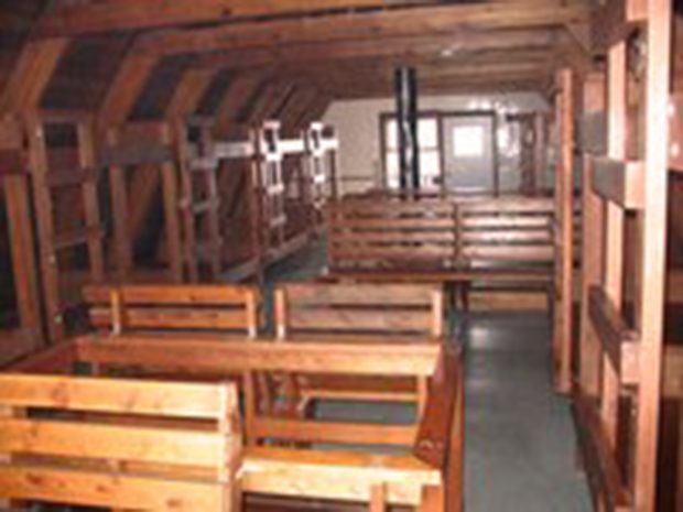 The wooden bunk beds line both sides of the second floor of the Hut. Down the middle are wooden benches with aisles on either side. The black smokestack of the wood stove is visible and stands in front of the white door on the far wall.