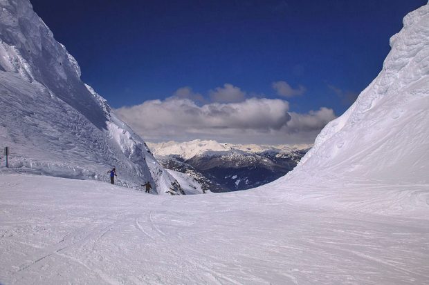 Two large snow walls line a steep ski run and sunshine reflects off the snow with deep blue sky above. Two skiers can be seen near one of the snow walls on the far left hand side of the ski run.