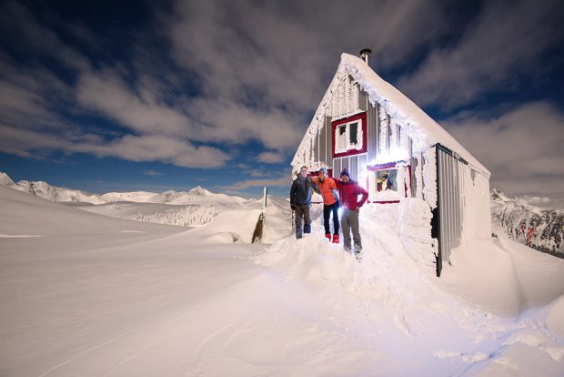A man wearing a dark blue jacket at grey pants, a woman wearing a bright orange jacket and black pants and a man wearing a toque, red jacket and light grey pants pose in front of the Hut built on Mount Brew.
