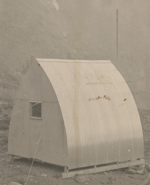Rear view of the corrugated metal siding, gothic arch hut with small square window installed at its final location. A large pole and steep rocky slope are in the background.