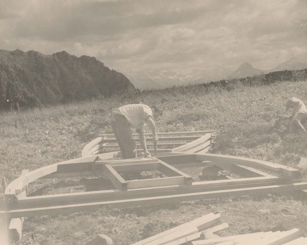 A man hunched over holding a hammer nailing in pieces to an end-wall. Construction has yet to begin on erecting the hut and grey clouds are visible in the background.