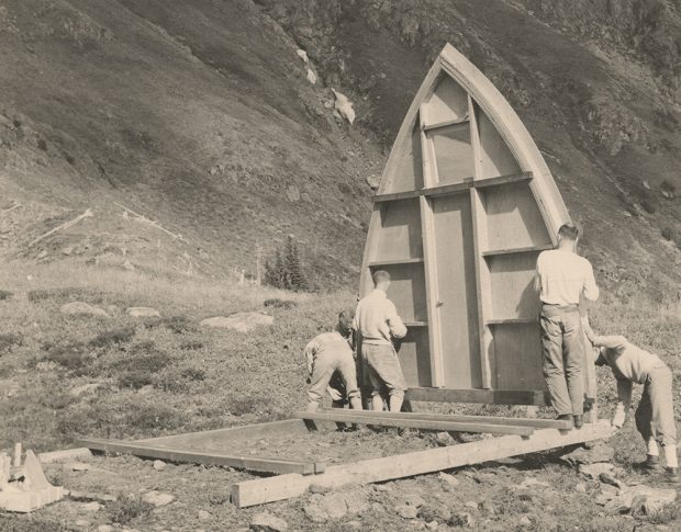 Two men stand holding one side of the arched front end-wall and three others work at attaching the other side to the foundation.