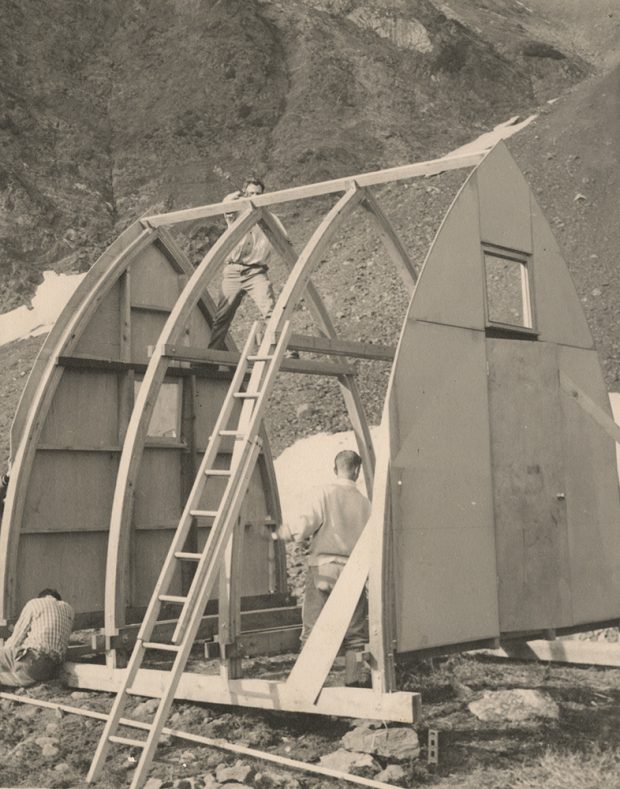 Three men are busy working on different aspects of the arched wooden frame of the Hut structure before roofing material can be installed.