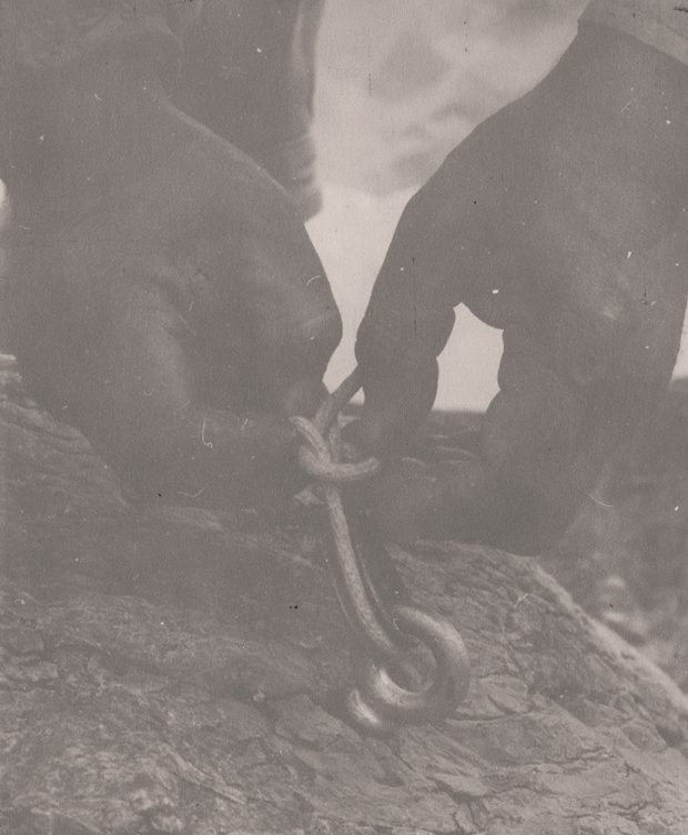 Close-up of hands tying knots. The cable has been looped through an eye-ring, which has been embedded into the bedrock located near the hut.