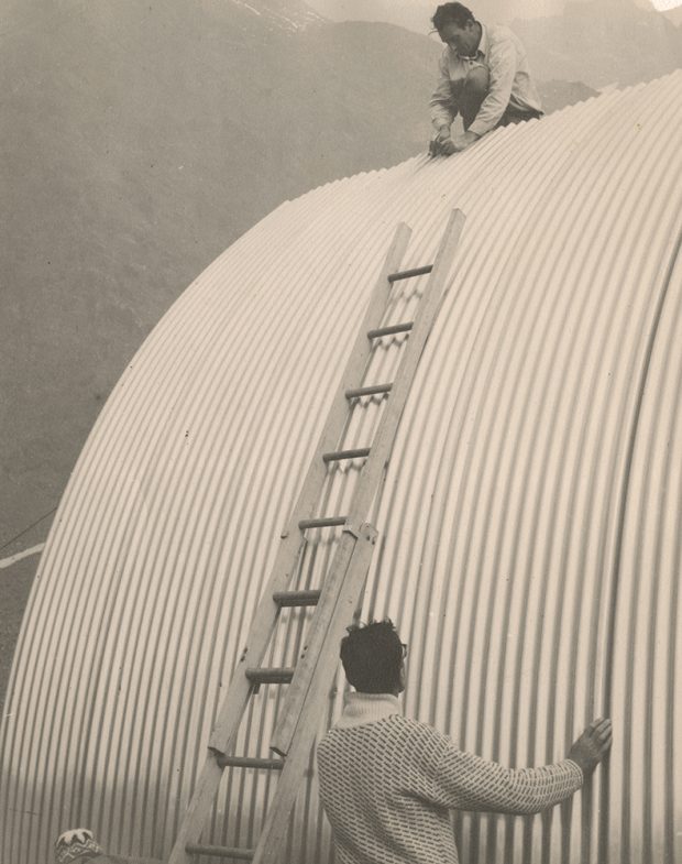 One man is perched at the top of the roof attaching a piece of siding and another man is holding the siding from the ground.