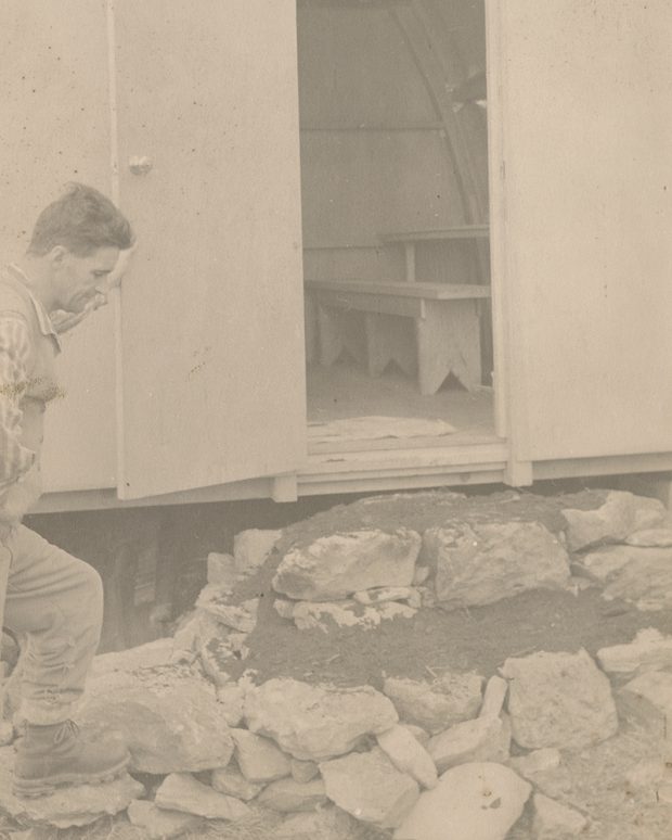 A man stands to the left holding on to the open door of the hut and is looking over the work he has done to the entryway.