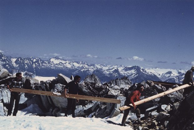 Two members of the work crew carry one long two-by-four and another stands holding one long board. Snow patches are on the ground and snowy peaks can be seen in the distance.