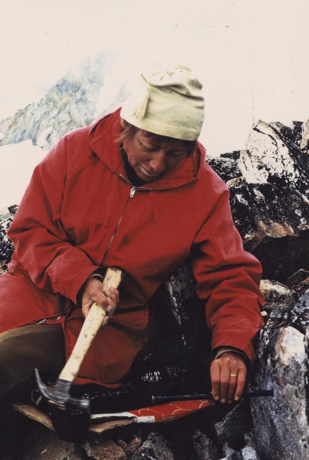 A woman in a red jacket and white toque sits leaned up against the rock while wielding a hammer and holding onto a long metal spike that is embedded in the bedrock.