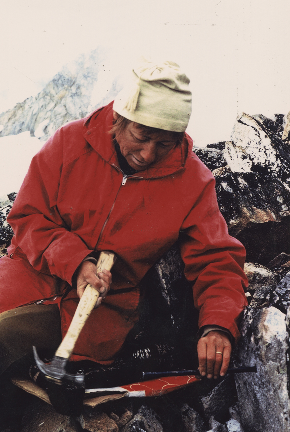A woman in a red jacket and white toque sits leaned up against the rock while wielding a hammer and holding onto a long metal spike that is embedded in the bedrock.
