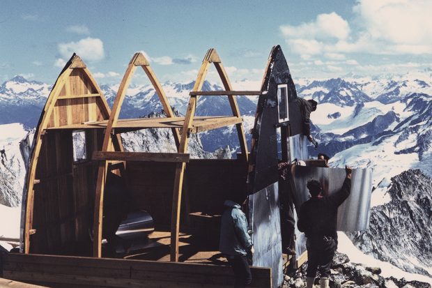 Construction on the Plummer Hut is underway. Men attach aluminum siding to one wall. A vast landscape of snowy mountains fills the background.