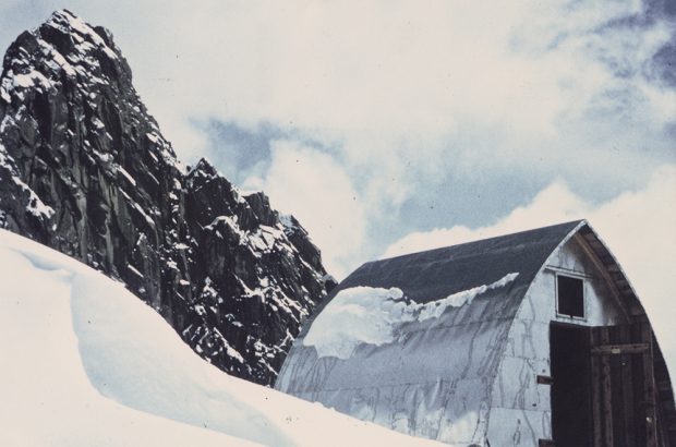 A rocky peak looms in the background with a light dusting of snow and the completed hut sits in the foreground surrounded by deep snow.