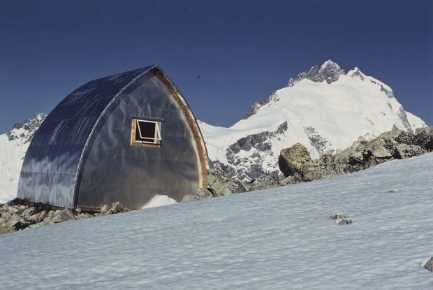 A thin layer of snow covers the slope on the backside of the completed Plummer Hut. The area around the hut has been completely cleared of snow. A large mountain peak covered in snow is in the background.