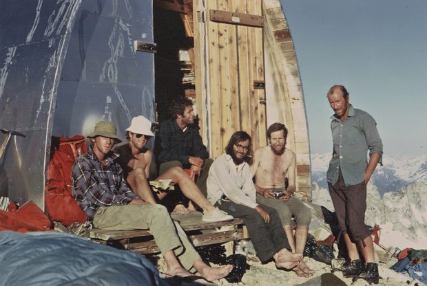 Two men in front wearing hats and 3 other men with out hats sitting on the front entryway of the Plummer Hut enjoying a break in the sunshine. One man is standing wearing a blue shirt and dark grey pants with snow-covered mountain peaks in the background.