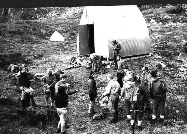 A group of students stand out front and to the right of the Batzer hut in the grassy alpine meadow.