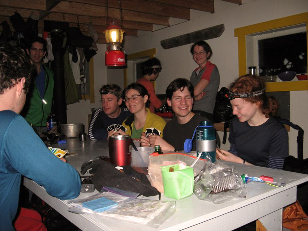 UBC-VOC Club trips have long held the tradition of playing card games. Club members are playing a game of UNO around the gray dining table inside the Hut. In the foreground, the right arm of a man wearing a blue shirt, holding his cards. A man in a green jacket stands at the far end of the table and a woman wearing a grey and red short sleeve shirt stands behind the 4 members sitting at the table. Another women in pink with her back turned is working away at the cooking station.