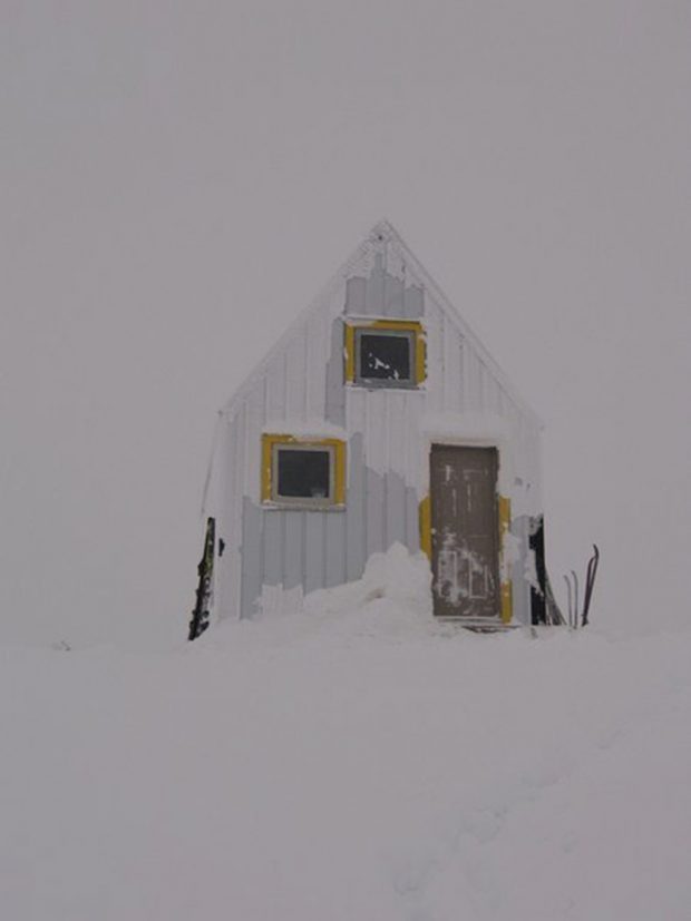 The grey sky looms behind the Hut. The yellow-framed windows and front door have been wind swept by snow.