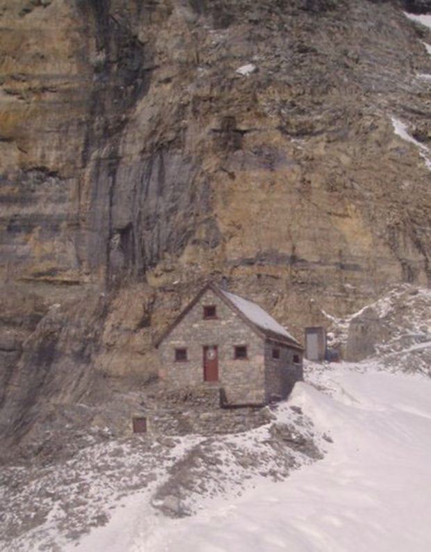 A stone shelter with an a-frame roof and a dark brown front door sits below a steep sedimentary rock face of the Rocky Mountains. In the background, the outhouse entrance is carved into the steep rock face and to the right of the Hut is a snow covered slope that runs to the edge of the image.