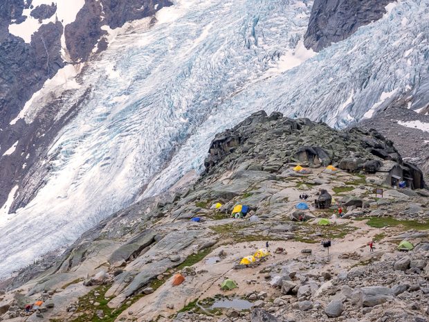 Photo looks down on a large open rock area dotted with tents located near a large glacier hanging on to a steep slope in the background.
