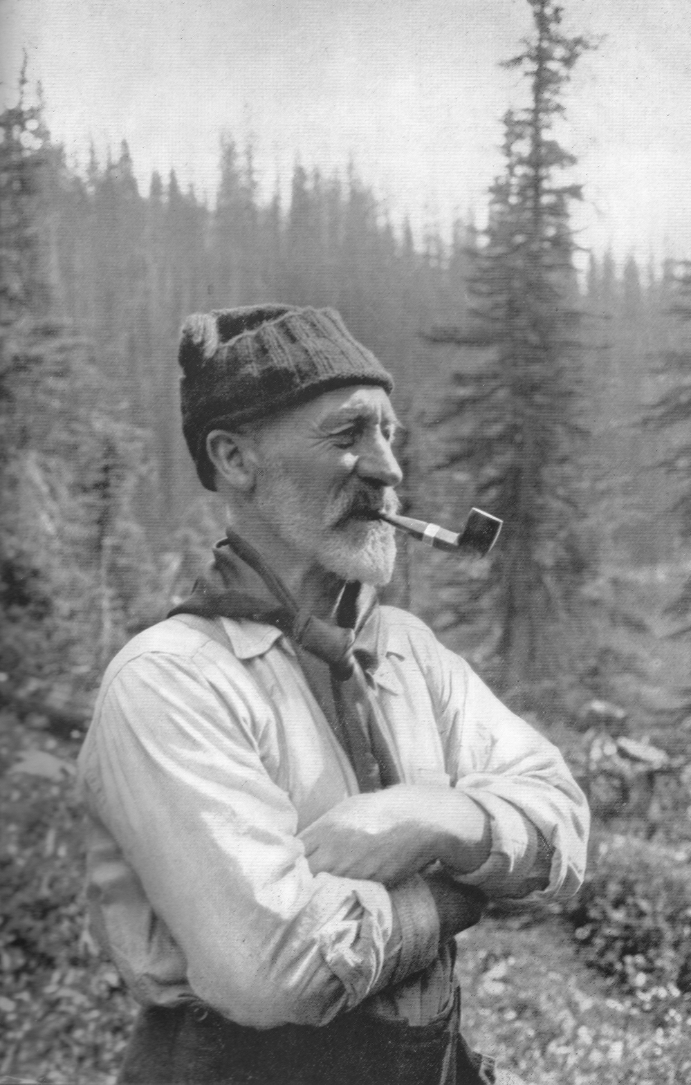 A black and white photograph of a bearded Arthur Wheeler with arms folded across his chest and pipe hanging out of his mouth, wearing a toque, scarf and a long-sleeve shirt with evergreen trees in the background.