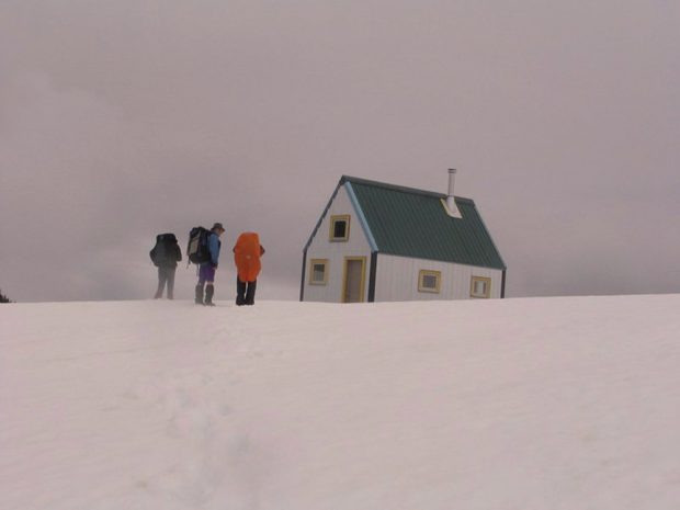 Three members stand wearing large backpacks on the snowy slope out front of the Hut. Grey clouds loom over the green roof of the Hut.