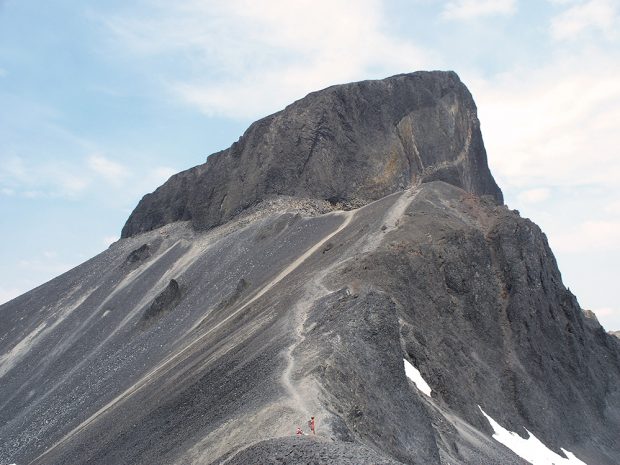 A close up of the Black volcanic rock face that makes the Tusk peak so easily identifiable. A person in an orange shirt stands along a ridge below the peak.