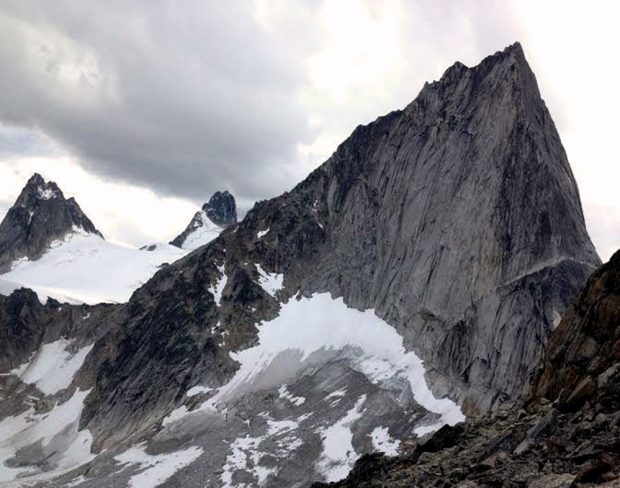The narrow grey spire tip with its jagged edged peak stands above a gravel snow covered slope. Two other grey spire peaks rise up from a glacier that sits next to the spired peak in the foreground.